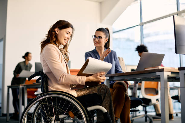 Happy disabled businesswoman and her colleague cooperating while working in the office. Happy businesswoman in wheelchair going through reports while talking to female coworker in the office. accessibility for persons with disabilities photos stock pictures, royalty-free photos & images