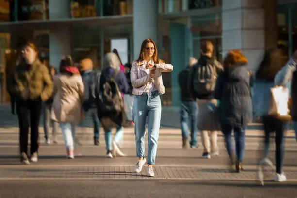 Photo of Young woman walking in the middle of crowded street and looking time at hand watches. Big city life.