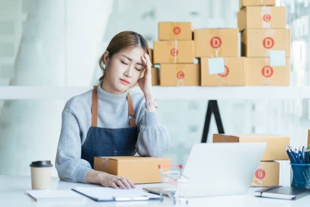 istock Young asian woman startup small business freelance holding parcel box and computer laptop and sitting on chair, getting headache. Online marketing packing box delivery concept 1368303474