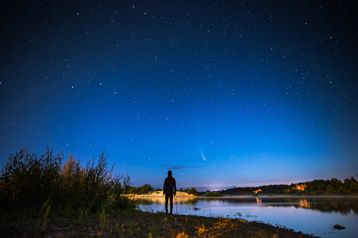 Man silhouette against background of starry sky. Comet in night sky. Summer starry sky. Beautiful landscape with stars. Long exposure. Fog over water. Atmospheric landscape. Morning twilight.