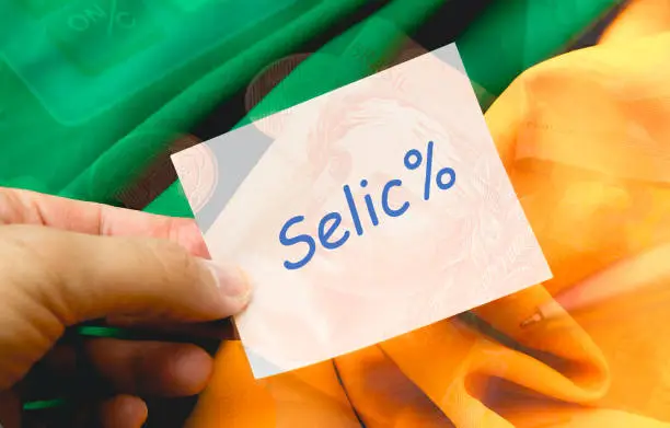 Photo of The word SELIC written on a note paper being held by a man. Background with the image of the Brazilian Real, money from Brazil, and with the colors green and yellow. Photo for Brazilian economy.