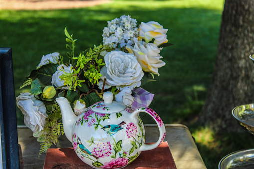 A beautiful teapot with brightly colored birds and flowers painted upon. And a vase of beautiful roses and white flowers.