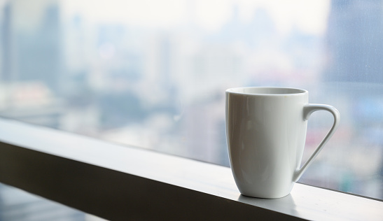 White mug of hot coffee near window with blurry of city view high building on background. Copy space
