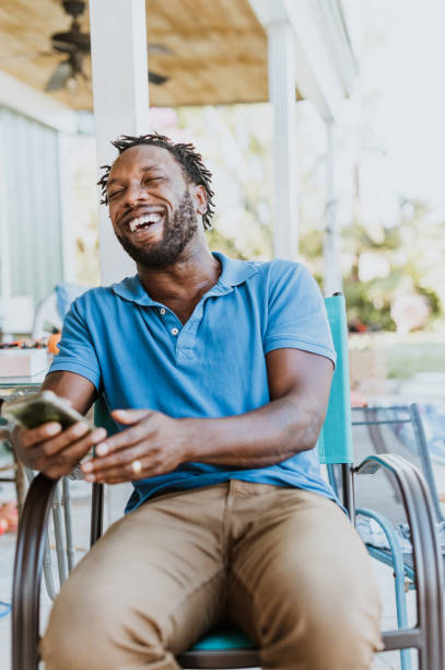 feliz chico negro sosteniendo el teléfono y riendo en un retrato casual al aire libre sincero - polo shirt african ethnicity men african descent fotografías e imágenes de stock