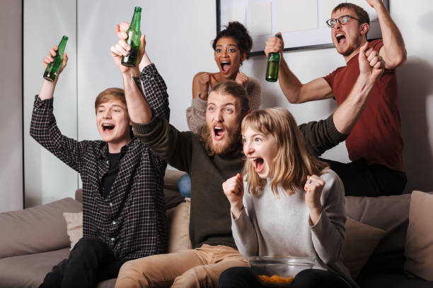 istock Joyful friends sitting on sofa happily screaming while watching tv with chips and beer at home 1368325782