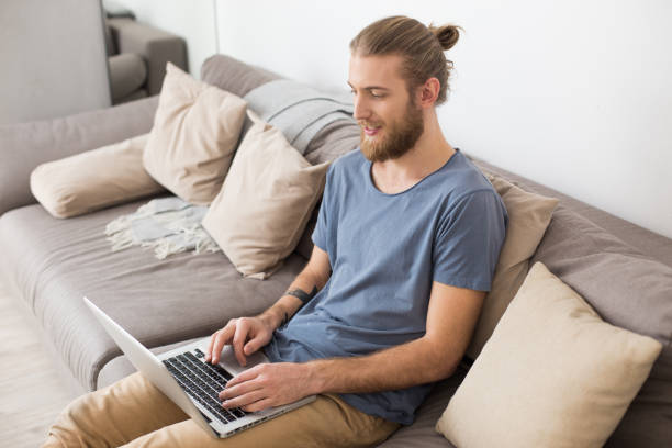 istock Young man sitting on big gray sofa and working on laptop at home 1368327549