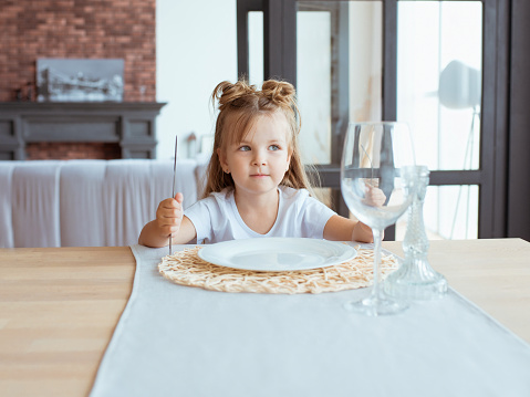 portrait of hungry little adorable girl in white t-shirt sitting at the table with fork, knife and empty plate in loft interior indoor