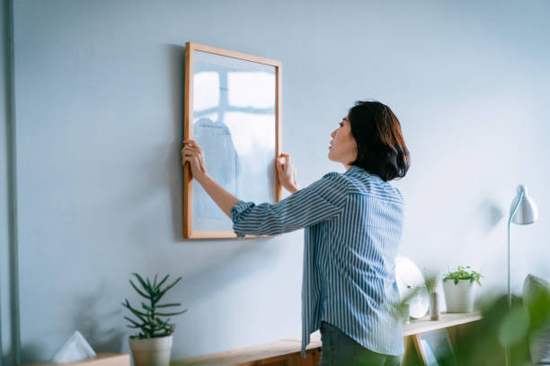 young asian woman decorating and putting up a picture frame on the wall at home - hangen stockfoto's en -beelden