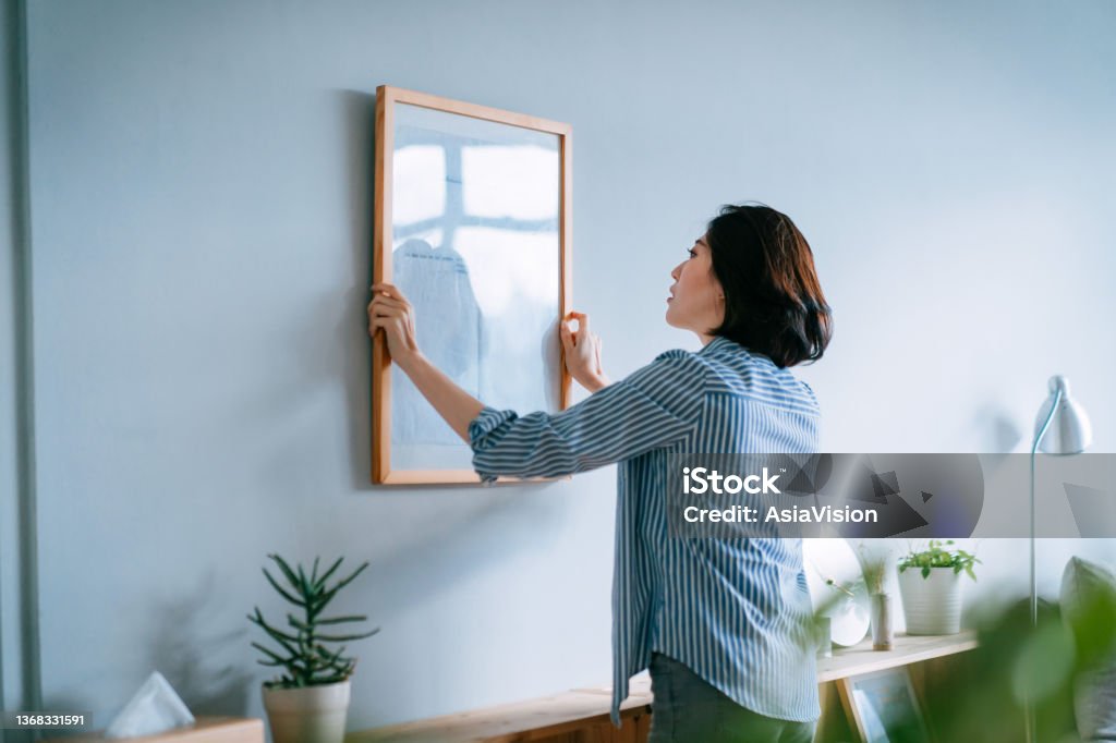 Young Asian woman decorating and putting up a picture frame on the wall at home Hanging Stock Photo