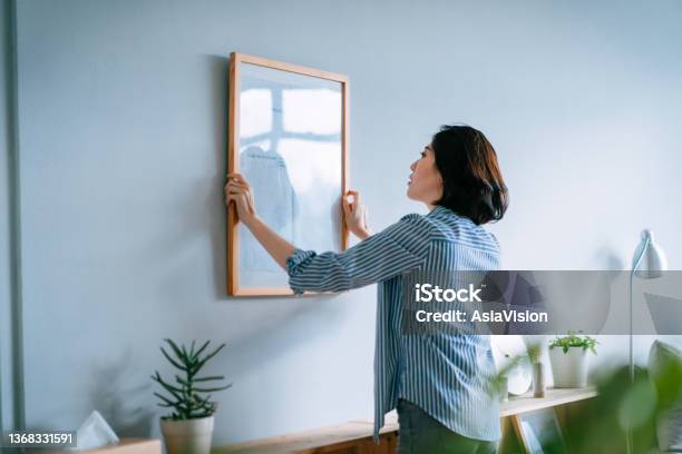 Young Asian Woman Decorating And Putting Up A Picture Frame On The Wall At Home Stock Photo - Download Image Now
