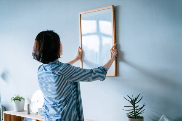 joven asiática decorando y colocando un marco de fotos en la pared de la casa - decorar fotografías e imágenes de stock