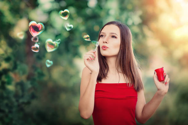 девушка в красном платье выдувает пузырьки воздуха в форме сердца. день святого валентина - sky sun grass tree стоковые фото и изображения