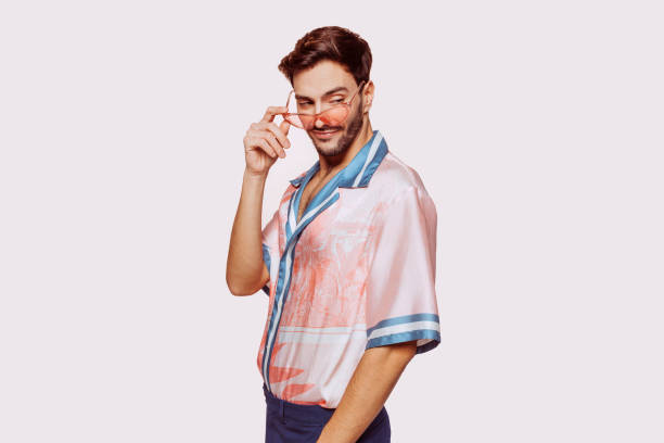 Young attractive caucasian gay man over beige wall smiling, posing and looking away at studio. Handsome homosexual in stylish clothes and pink sunglasses.
