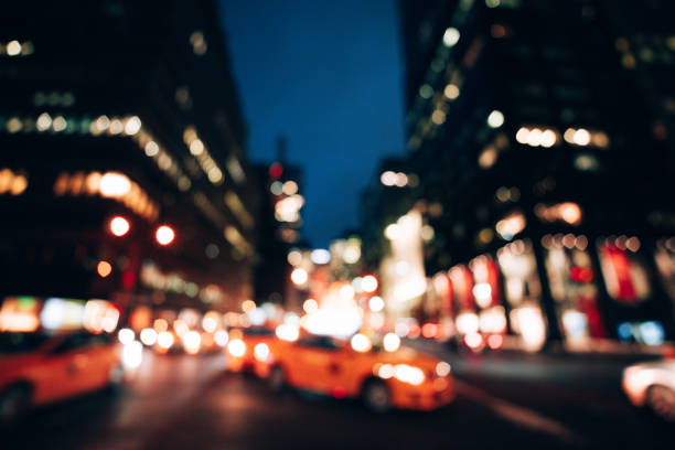 Defocused, zoomed image of a downtown city street at night. Unique photographic effects create a blurred motion vanishing point. Red, orange and yellow hues with great copyspace at bottom. A red vehicle in center of image driving down the busy road. Bright lights in background. Taillights, headlights.