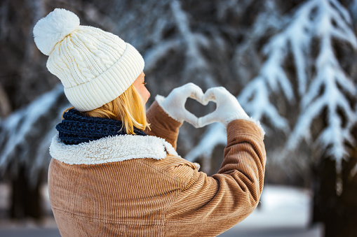 Blonde beautiful young woman smiling wearing winter jacket on the winter day, forming a heart shape with her fingers