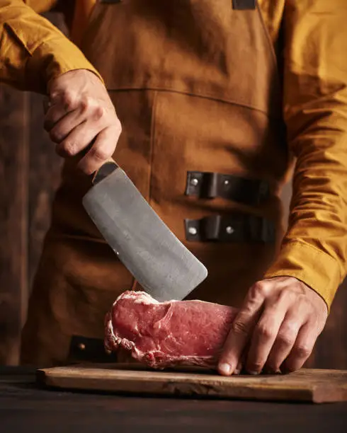 A butcher in a leather apron cuts a piece of meat with a large knife.