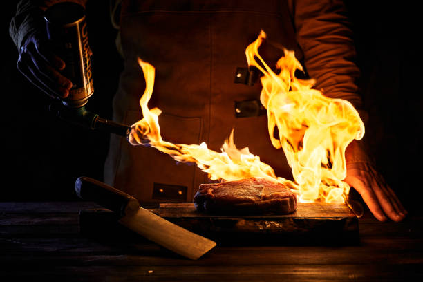 A butcher with a burner burns a steak. A piece of raw meat with fire.