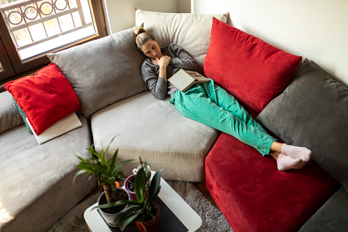 Tired young woman lying on sofa in living room and sleeping with book on her body
