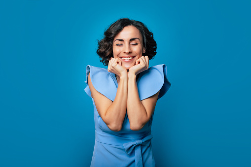 Close up portrait of an excited happy gorgeous girl in a long blue dress, who is looking in the camera while she posing with bright toothy smile and isolated on blue background