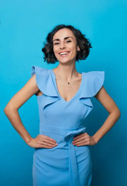 Photo of Vertical photo of a charming smiling woman in a blue dress, who is looking in the camera while standing and posing isolated on blue background