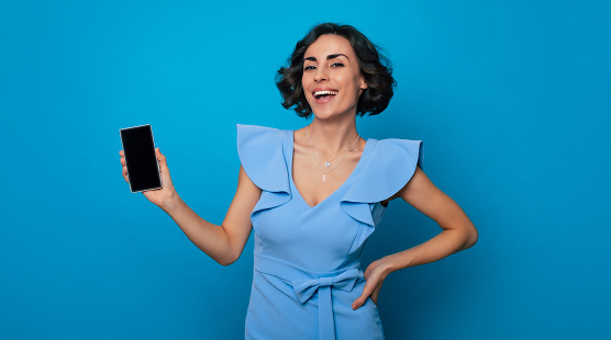 Fashionable beautiful smiling woman in blue dress shows a smart phone with blank screen on the camera and isolated on blue background