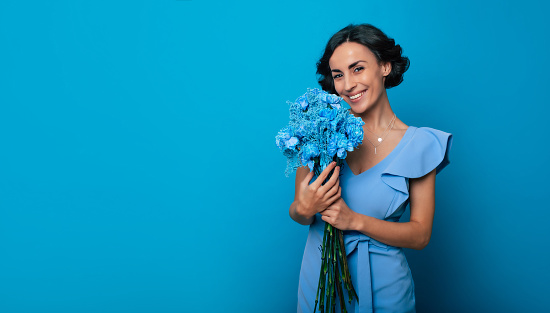 Young gorgeous woman in a bright blue dress is looking in the camera with a big smile, holding a bunch of blue flowers in her hands. International Women's Day