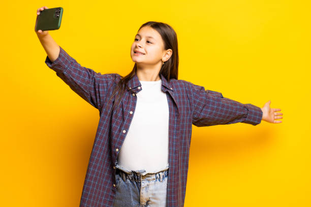 istock Little kid gir over yellow background holds modern mobile phone and makes video call waves palm in hello gesture. People modern technology concept 1368376827