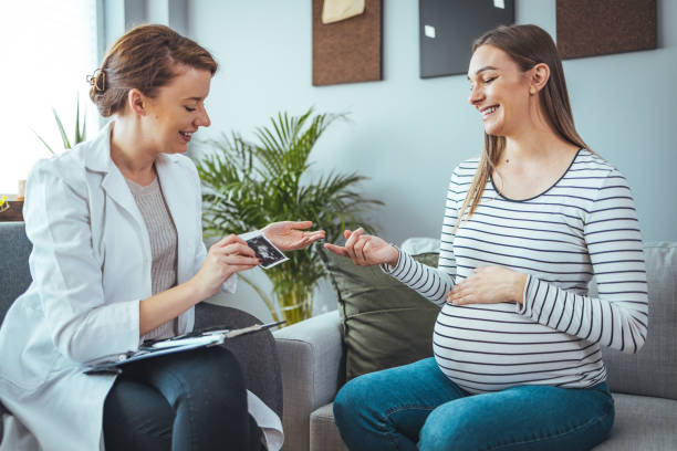 Young  woman smiles while looking at her baby's ultrasound image. Young  woman smiles while looking at her baby's ultrasound image. She is meeting with a home healthcare nurse. Pregnant woman and doctor looking at her baby ultrasound picture Midwife stock pictures, royalty-free photos & images