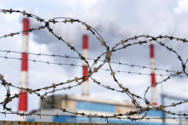 barbed wire at a thermal power plant, in the background a blurred background of three white-red pipes with the release of a large amount of smoke and steam against the blue sky in winter barbed wire at a thermal power plant, in the background a blurred background of three white-red pipes with the release of a large amount of smoke and steam against the blue sky in winter. barbed wire wire factory sky stock pictures, royalty-free photos & images