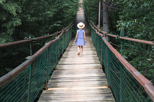 A little girl in a straw hat walks on the old suspension bridge