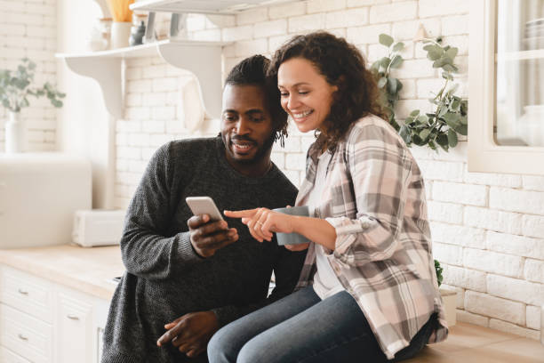 中年の成熟した幸せなアフリカのカップルは、ソーシャルメディアサーフィン、写真の共有、電子メールのチェック、自宅のキッチンでリラックスしたモバイルアプリケーションを使用して� - sharing mobile phone application software e mail ストックフォトと画像