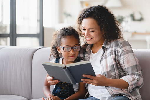 Madre madre afroamericana leyendo cuentos de hadas libro novela con hija niña, aprendiendo juntos, ayudando con la tarea para la escuela. La tutora enseña a la colegiala en casa. Educación en el hogar photo