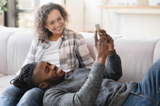中年の成熟した幸せなアフリカのカップルは、ソーシャルメディアサーフィン、写真の共有、電子メールのチェック、ソファで自宅でリラックスしたモバイルアプリケーションを使用してス� - sharing mobile phone application software e mail ストックフォトと画像
