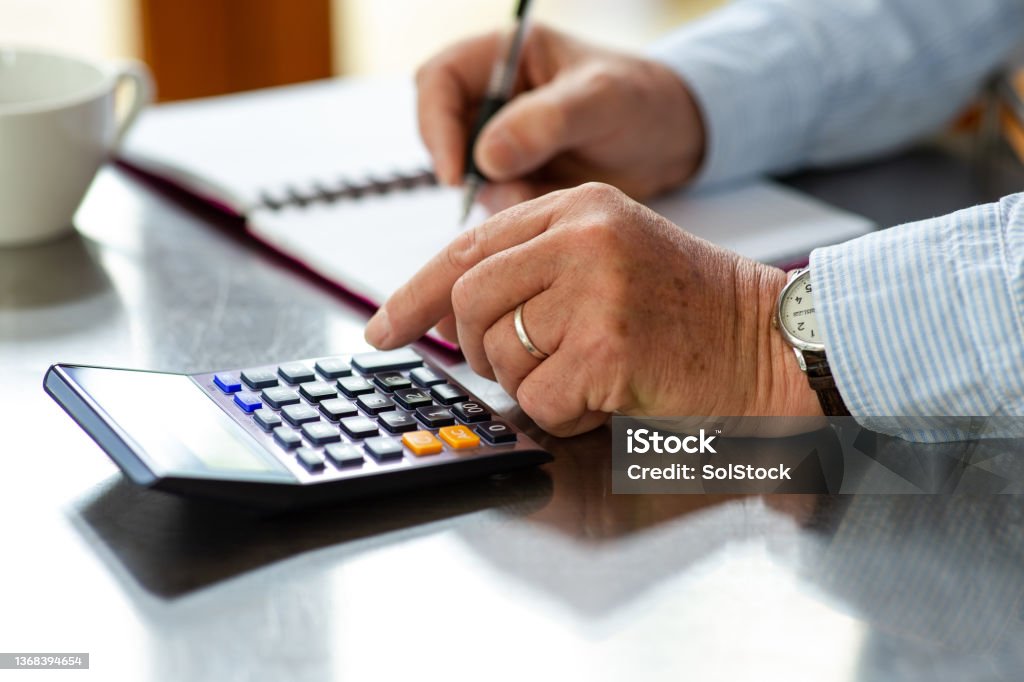 Calculating His Monthly Spend An unrecognisable businessman using a calculator and writing in a notepad at home on a counter. He is updating his personal finances and working out bills. Home Finances Stock Photo