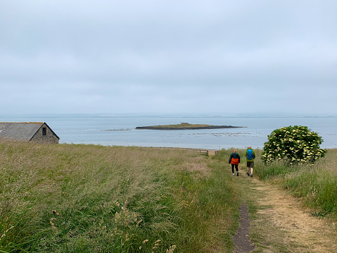 A male and female friend walking on a path through a grass area at Holy Island, Northumberland with the coastline in front of them. They are enjoying a day out together.