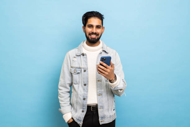 istock Young happy bearded man posing isolated over blue wall background using mobile phone chatting. 1368396007