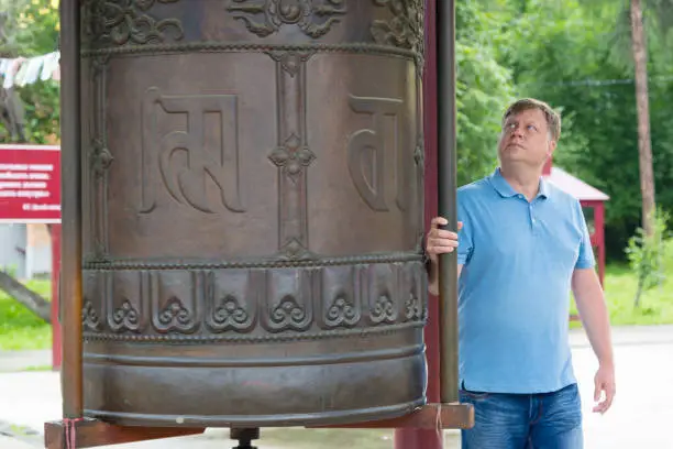 Photo of An adult tall man turns a metal drum in a Buddhist temple with his head down