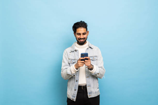 istock Young happy bearded man posing isolated over blue wall background using mobile phone chatting. 1368397741