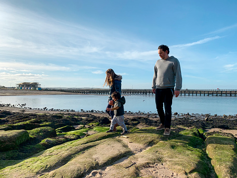 A man with his son and niece, walking over rocks on the beach at Amble, Northumberland. The girl is holding hands with her cousin, helping him walk over the rocks.