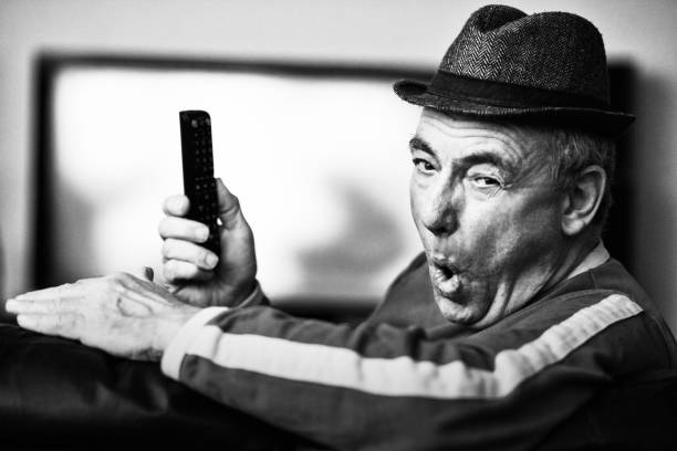 Senior man in his late 60s holds TV remote and looks back at you with an expression that could be interpreted several ways.
