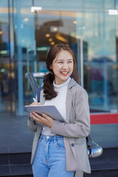 Asian woman holding a tablet with a bag standing in front of a shopping mall selling work-studying ideas outdoors. Asian woman holding a tablet with a bag standing in front of a shopping mall selling work-studying ideas outdoors. asian adult student stock pictures, royalty-free photos & images