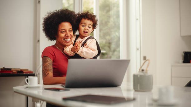 美しい若いママは、アパートのリビングルームでラップトップコンピュータを使用して、彼女の素敵な赤ちゃんの息子を保持しています。母は自宅、オンラインショッピング、ソーシャルメ� - family single mother black mother ストックフォトと画像