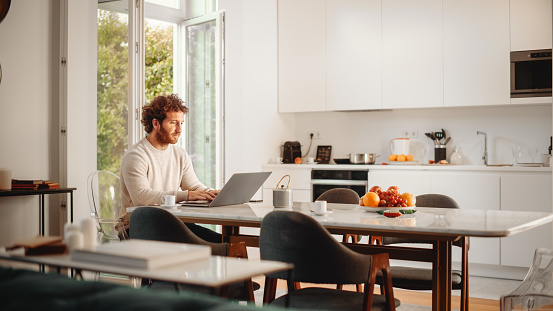 Handsome Adult Man with Ginger Curly Hair Using Laptop Computer, Sitting in Living Room in Apartment. Male Drinks Espresso, Working from Home, Online Shopping, Watching Videos or Writing Emails.