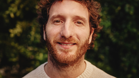 Close Up Portrait of a Happy Young Adult Male with, Brown Eyes, Curly Ginger Hair and Beard Posing for Camera. Handsome Diverse Caucasian Male Smiling on Green Nature Background.
