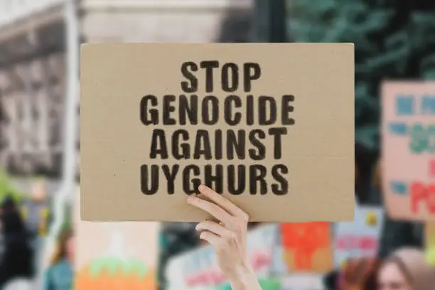 The phrase " Stop genocide against uyghurs " on a banner in men's hand. Human holds a cardboard with an inscription. Communism. Totalitarianism. Kill. Prison. Colonies. Control. Discrimination. Rights