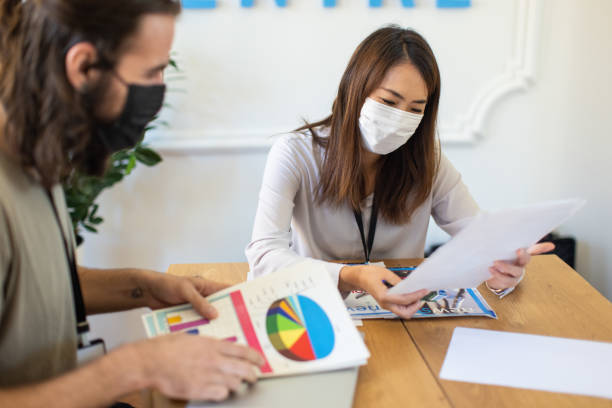 istock Female Japanese editor, with young male journalist talking about documents, while wearing protective face mask 1368403391