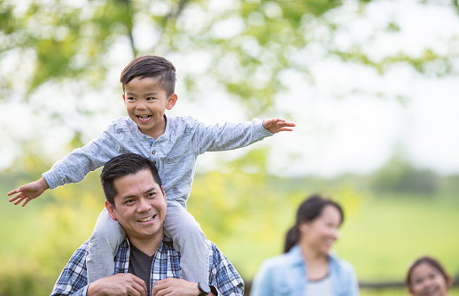 A young Father of Filipino decent, holds his son on his shoulders as he carries him on a family walk together.  The young boy has his arms stretched out as he pretends to fly through the air, and the Mother and her daughter can be seen walking in the background.  They are all dressed casually and smiling as they enjoy the sunny fall day and their time together.