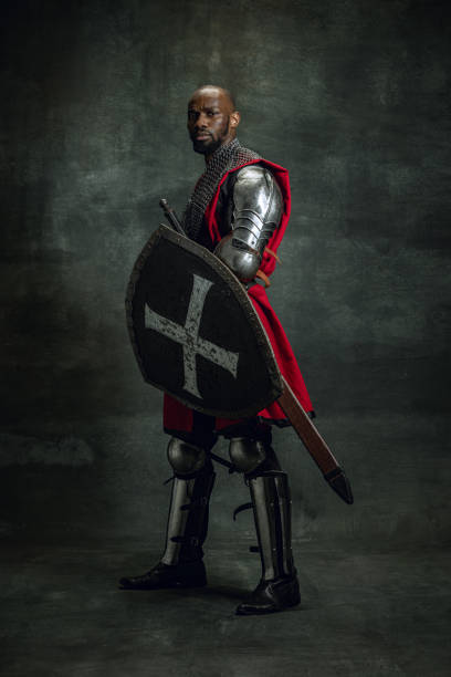 Vintage style portrait of brutal dark skinned man, medieval warrior or knight with wounded face wearing armour isolated over dark background. Comparison of eras, history Vintage style portrait of brutal dark skinned man, medieval warrior or knight with wounded face wearing armour isolated over dark background. Comparison of eras, history, renaissance style knight person stock pictures, royalty-free photos & images