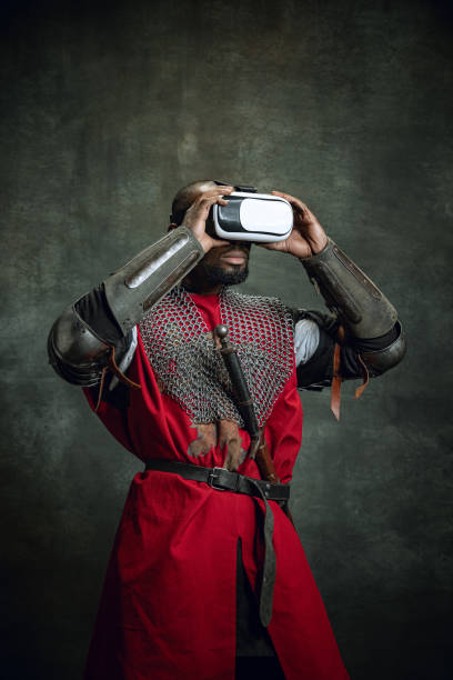 Modern gadgets. One dark skinned man, medieval warrior or knight using VR headsets isolated over dark vintage background. Comparison of eras, history, technology Modern gadgets, new games. One dark skinned man, medieval warrior or knight using VR headsets isolated over dark vintage background. Comparison of eras, history, renaissance style black knight stock pictures, royalty-free photos & images