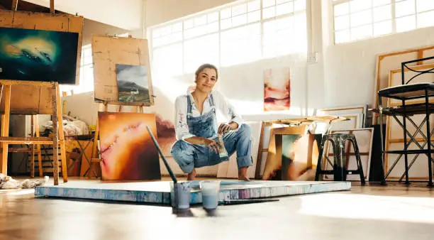 Creative painter squatting close to her painting in an art studio. Artistic young woman working on a new painting on the floor. Cheerful young artist looking at the camera while holding a paintbrush.